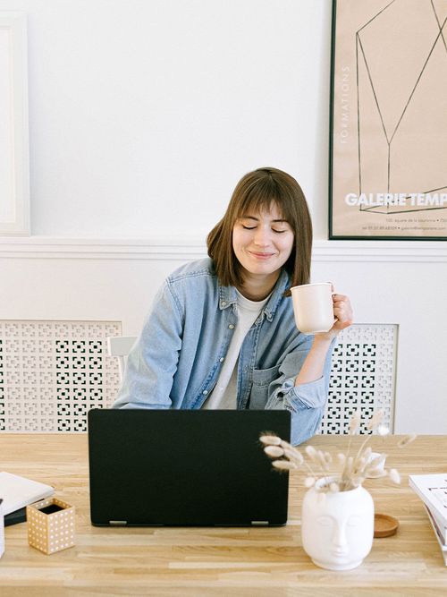 Frau sitzts an Holztisch, hält eine Tasse in der Hand und schaut auf ihren Laptop.