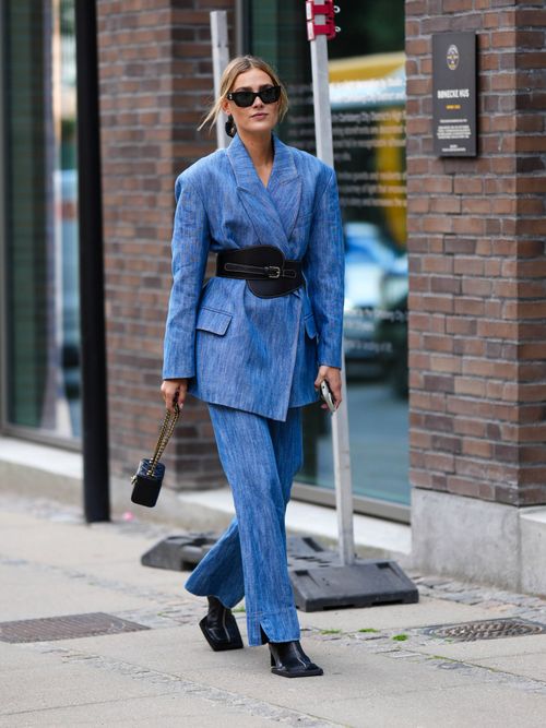 Street Style Look von der Copenhagen Fashion Week, wo ein Gast einen Jeans-Hosenanzug mit breitem Taillengürtel, Boots und Sonnenbrillen trägt.