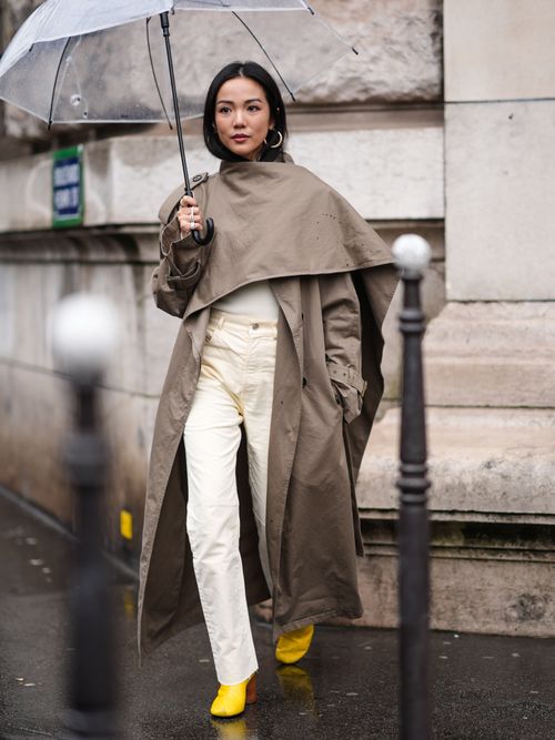 Yoyo Cao trägt auf der Paris Fashion Week einen Trenchcoat, beige Hose, gelbe Stiefeletten und hält einen transparenten Regenschirm in der Hand.
