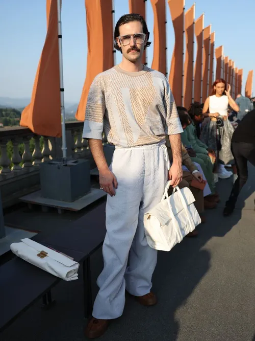 Nelson Tiberghien trägt ein sommerlich gemustertes T-Shirt, einer weißen Hose, Sonnenbrille und einer Handtasche auf der Milan Fashion Week