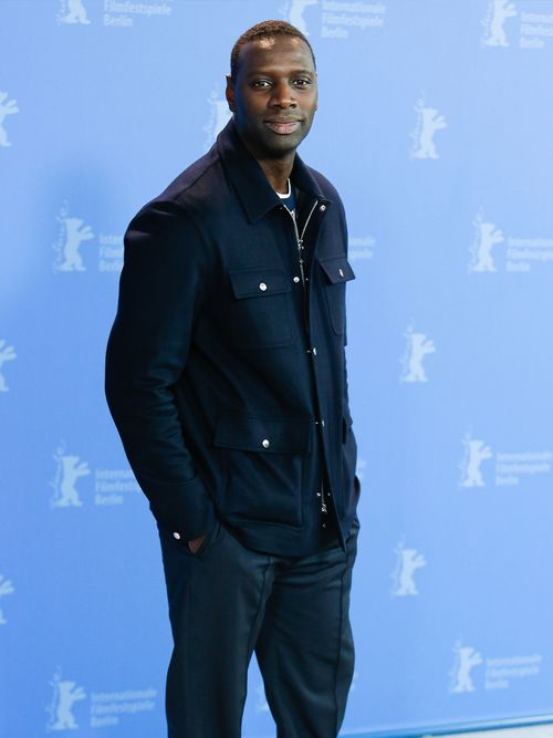 Omar Sy in einer blauen Jacke vor einem hellblauen Hintergrund.