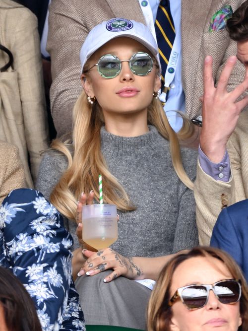 Ariana Grande is seen at the Wimbledon Tennis Championships 2023 wearing a white Wimbledon baseball hat, round green sunglasses, a sparkly grey short-sleeved jumper, and grey wool trousers.