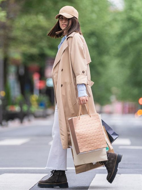 Street Style Foto von einer Frau im Trenchcoat und Capy, mit weißer Hose, die viele Sale Shopping Taschen trägt.