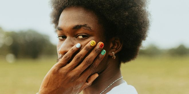 Mann mit Afro trägt ein weißes T-Shirt und hat bunt lackierte Fingernägel.