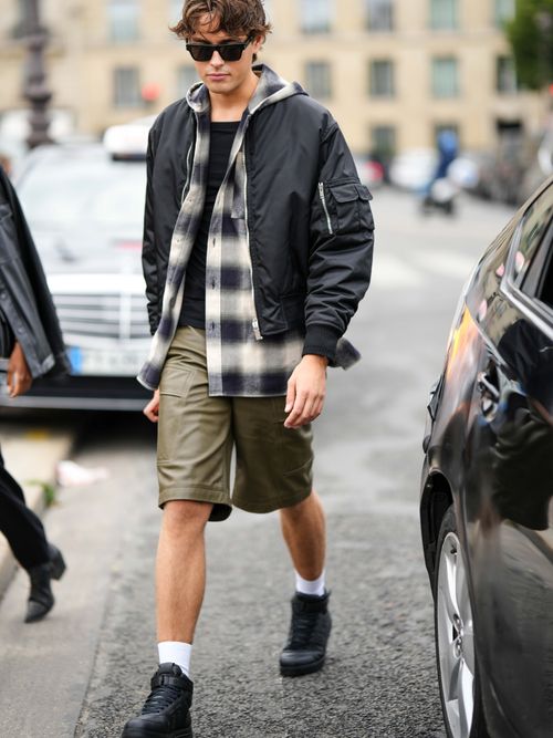 Street Style Look von der Paris Fashion Week, wo ein Gast eine schwarze Bomberjacke, kariertes Hemd, schwarzes T-Shirt, grüne Bermudas und schwarze Sneakers trägt.