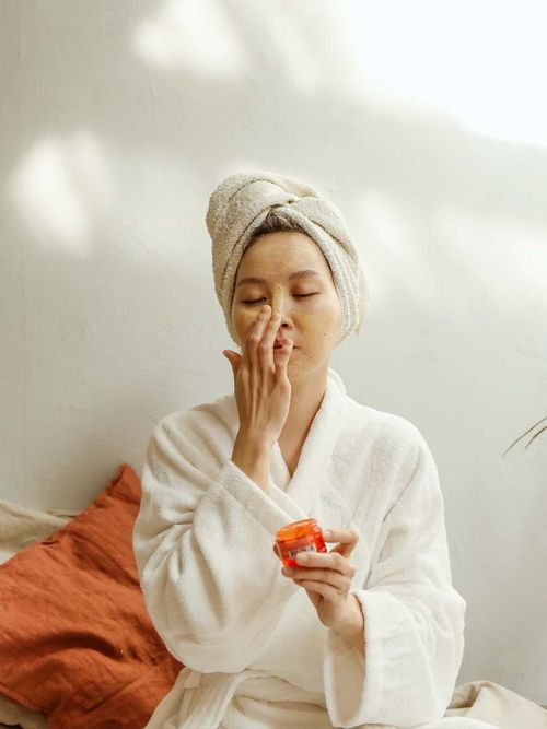 Frau im Bademantel und mit Handtuchturban trägt gerade eine Gesichtsmaske auf.