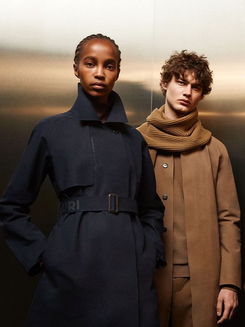 Eine Frau in einem schwarzen Trenchcoat und ein Mann in einem beigen Mantel von Alphatauri.