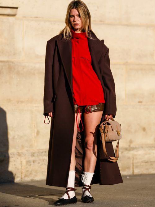 Natalia Verza trägt einen braunen Mantel, eine rote Jacke, Ledershorts, weiße Socken und schwarze Ballerinas in Paris.
