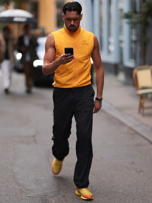 Street Style Look von der Copenahgen Fashion Week, wo ein Gast ein orangefarbenes Daunen-Gilet, schwarze Trainingshose und orangefarbene Sneaker trägt.