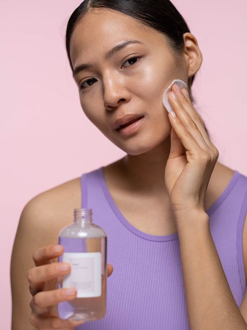 Frau mit lila Top hält ein Gesichtswasser in einer Hand und mit der anderen wischt sie mit einem Wattepad über ihr Gesicht.