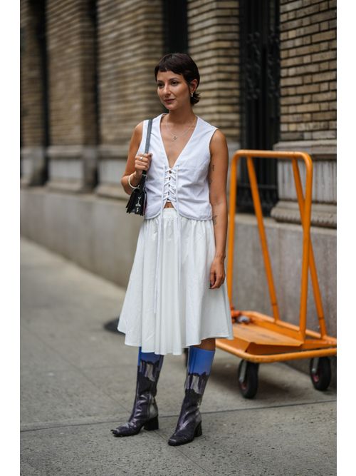 Alyssa Coscarelli wears silver earrings, a silver chain pendant necklace, a white laces V-neck / sleeveless top, a black shiny varnished leather shoulder bag, silver bracelets, silver rings, a white pleated knees skirt, black and blue gradient print pattern shiny varnished leather knees boots.