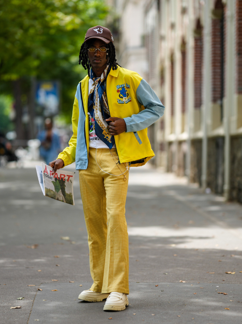 Street Style Look von der Paris Fashion Week, wo ein Mann eine gelbe Bomberjacke mit Halstuch und gelber Boot-Cut Hose mit Capy trägt.