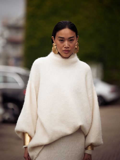 Street Style Look von der Copenhagen Fashion Week, wo ein Gast einen weißen Rollkragenpulli, beigen Strickrock und Goldschmuck trägt.