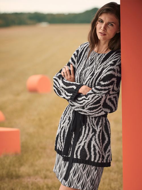 Frau mit gestrickten Cardigan und Rock lehnt an einer orangenen Wand auf einer Wiese.