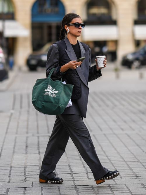 A guest wears black sunglasses, a white pearl earrings, a black t-shirt, a gray oversized blazer jacket, a black quilted sleeveless gilet, dark gray suit pants, a dark green with embroidered white print pattern handbag, black shiny leather nailed / studded with brown clogs outside Schiaparelli, during Paris Fashion Week.